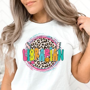May include: White t-shirt with a colorful graphic design that says "LIBRARIAN" in a circle with a leopard print background. The design also includes stars, lightning bolts, and a smiley face.