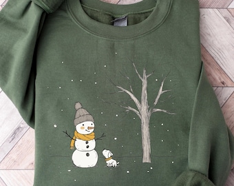 May include: A green sweatshirt with a snowman and a dog in a snowy scene. The snowman is wearing a brown hat and a yellow scarf. The dog is white with a brown spot on its back.