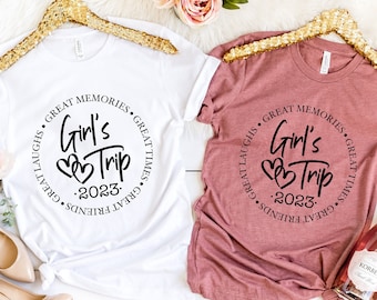 May include: Two white t-shirts with a black circle design that says "Girl's Trip 2023" with hearts. One shirt is white and the other is a light pink color. The shirts are hanging on gold hangers.