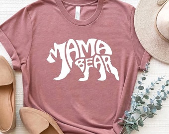 May include: A mauve t-shirt with a white bear silhouette design that says "Mama Bear". The shirt is laid out on a white surface with a brown hat, two brown shoes, and a sprig of eucalyptus.