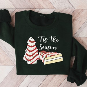 May include: A dark green sweatshirt with a white graphic of a Christmas tree and a slice of cake. The text "'Tis the Season" is printed in white on the sweatshirt.
