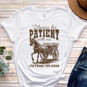 Puede incluir: Camiseta blanca con un gráfico marrón de un caballo tirando de un carro. El texto en la camiseta dice "Please be PATIENT with me I'M FROM THE 1900s".