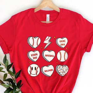 May include: Red t-shirt with a baseball-themed design. The design features baseballs, hearts, and phrases like "Game Day", "Kinda Day", and "Ballpark".