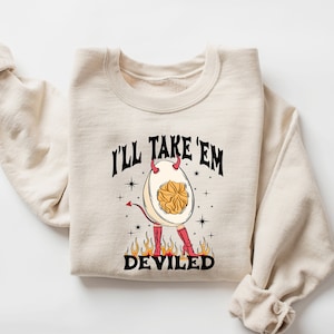 May include: A beige sweatshirt with the text "I'll take 'em deviled" printed on the front. The text is in black and there is a cartoon image of a deviled egg with horns and red boots.