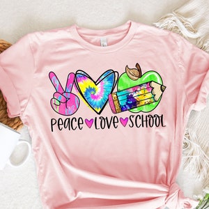 May include: Pink t-shirt with a peace sign, tie-dye heart, and apple with a pencil. The text "Peace Love School" is printed below the design.