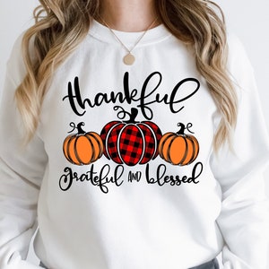 Puede incluir: Sudadera blanca con el texto "Thankful Grateful and Blessed" en letras negras. Se muestran tres calabazas, una con un patr&oacute;n de cuadros rojos y negros y dos con tallos naranjas y verdes.