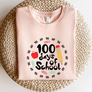 May include: A pink t-shirt with a graphic design that says "100 days of school" with a red apple, a pencil, and paper clips around the text.