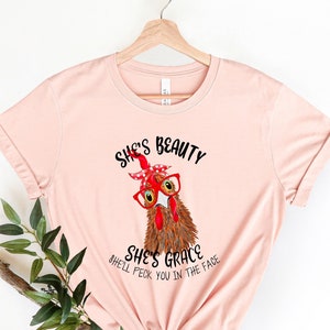 May include: A pink t-shirt with a graphic of a chicken wearing red glasses and a red bandana. The text on the shirt reads "She's Beauty She's Grace She'll Peck You In The Face."