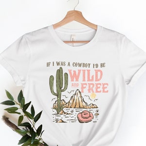 May include: White t-shirt with a graphic print of a cactus, mountains, and a cowboy hat. The text reads "If I was a cowboy I'd be wild and free".