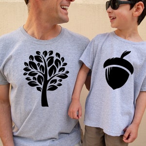 May include: Two people wearing matching gray t-shirts with black silhouettes. The adult shirt features a tree design, while the child's shirt has an acorn design.