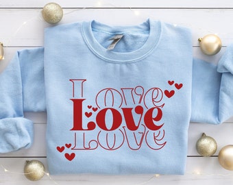 May include: A light blue sweatshirt with the word "LOVE" in red, stylized text. Small red hearts are scattered around the word. The sweatshirt is displayed on a white wooden surface with gold ornaments.