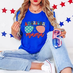Puede incluir: Una mujer con una camiseta azul con el texto "American Mama" y unas gafas de sol con un dise&ntilde;o de bandera estadounidense. Est&aacute; sosteniendo unas gafas de sol rojas, blancas y azules en su mano derecha.