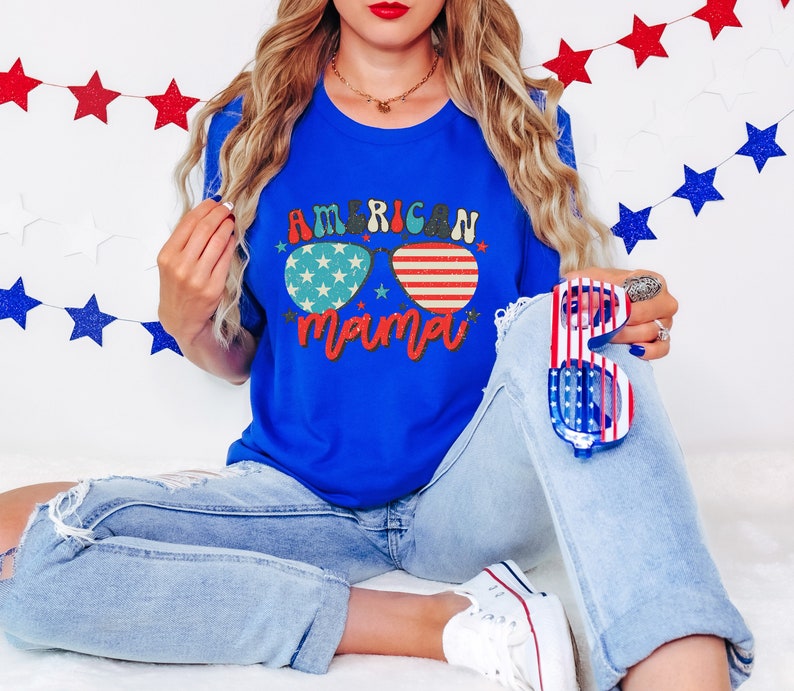 Puede incluir: Una mujer con una camiseta azul con el texto "American Mama" y unas gafas de sol con un dise&ntilde;o de bandera estadounidense. Est&aacute; sosteniendo unas gafas de sol rojas, blancas y azules en su mano derecha.