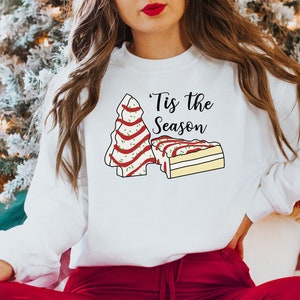 May include: White sweatshirt with a Christmas tree and a slice of cake design. The text "'Tis the Season" is printed on the sweatshirt.