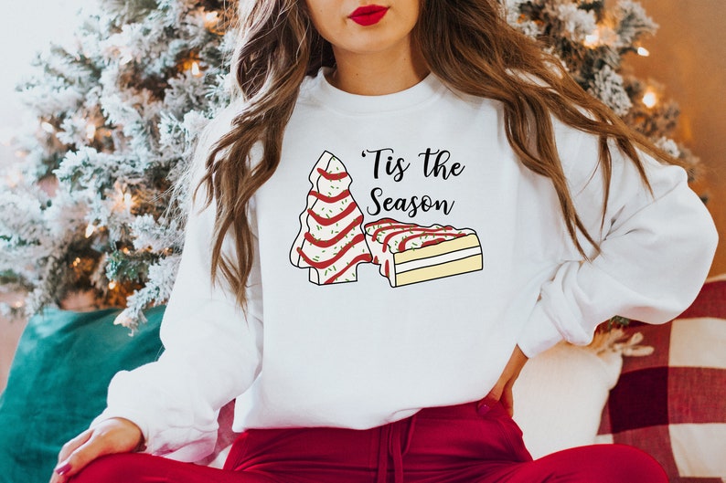 May include: White sweatshirt with a Christmas tree and a slice of cake design. The text "'Tis the Season" is printed on the sweatshirt.