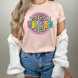 May include: A pink t-shirt with a colorful graphic design that says "LIBRARIAN" in a circle with a leopard print background. The graphic also includes stars and lightning bolts.