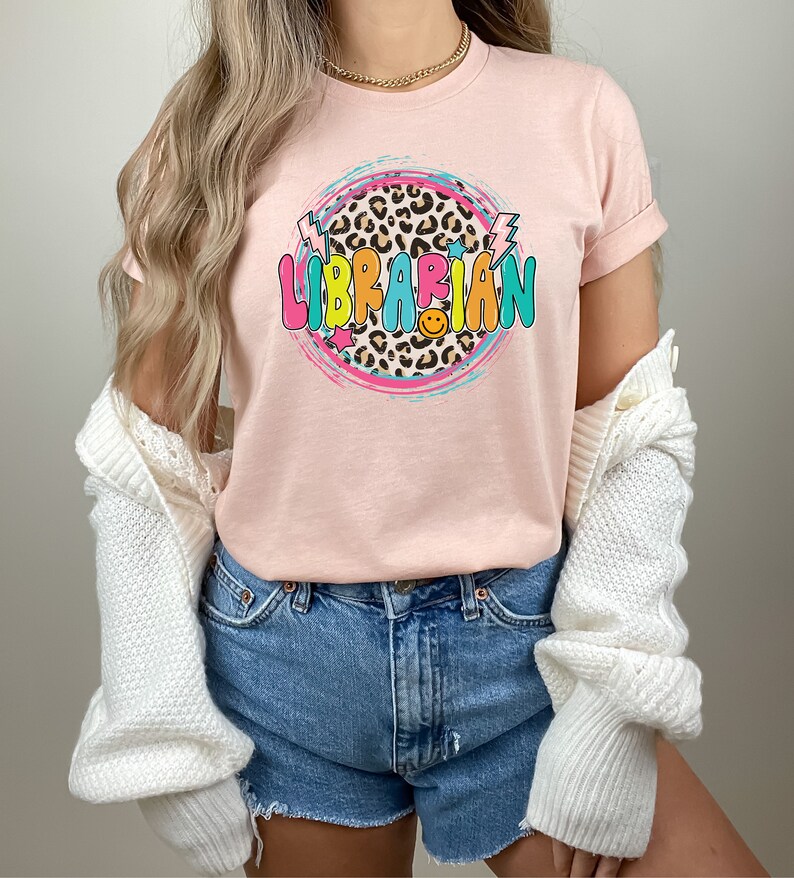 May include: A pink t-shirt with a colorful graphic design that says "LIBRARIAN" in a circle with a leopard print background. The graphic also includes stars and lightning bolts.