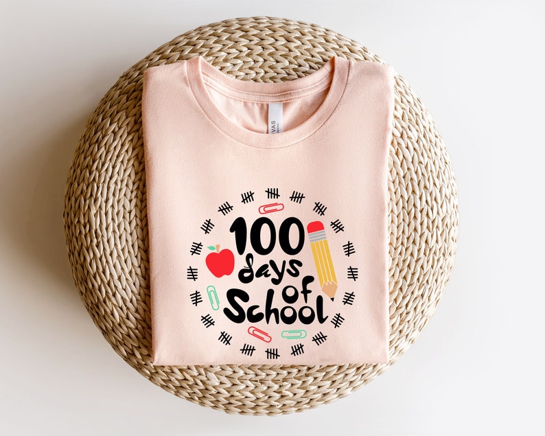 May include: A pink t-shirt with a graphic design that says "100 days of school" with a red apple, a pencil, and paper clips around the text.
