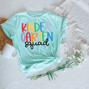 May include: A light blue t-shirt with the text "kindergarten squad" in rainbow colors.