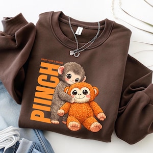 May include: A brown sweatshirt featuring a cartoon illustration of two monkeys. One monkey is hugging an orange plush monkey. The word "Pinch" is printed vertically in orange, with "Baby Love's Mama" above it. A silver necklace is draped on the sweatshirt.