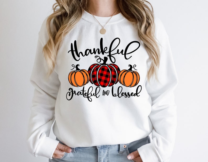 Puede incluir: Sudadera blanca con el texto "Thankful Grateful and Blessed" en letras negras. Se muestran tres calabazas, una con un patr&oacute;n de cuadros rojos y negros y dos con tallos naranjas y verdes.
