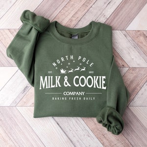 Puede incluir: Una sudadera verde con un dise&ntilde;o gr&aacute;fico blanco que dice "North Pole Milk & Cookie Company Baking Fresh Daily" con una ilustraci&oacute;n de Santa Claus y renos.