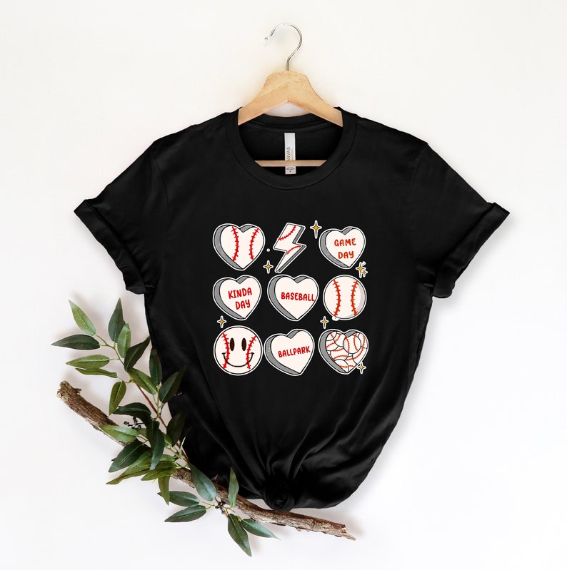 May include: Black t-shirt with a baseball theme. The shirt features a variety of baseball-related graphics, including baseballs, bats, and hearts with baseball-themed text. The text on the hearts includes "Game Day", "Kinda Day", "Baseball", "Ballpark".