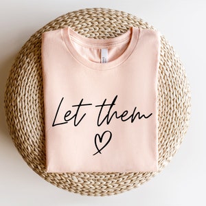 K&ouml;nnte beinhalten: Ein pfirsichfarbenes T-Shirt mit dem Text "Let them &hearts;" in schwarzen Buchstaben.
