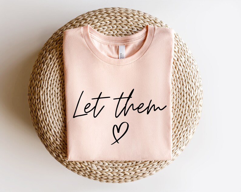 K&ouml;nnte beinhalten: Ein pfirsichfarbenes T-Shirt mit dem Text "Let them &hearts;" in schwarzen Buchstaben.
