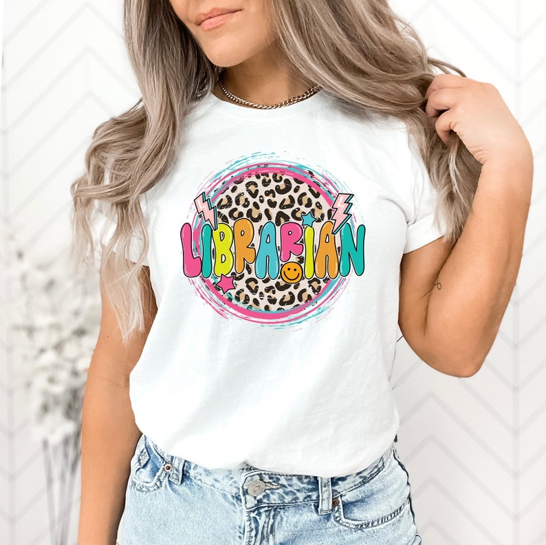 May include: White t-shirt with a colorful graphic design that says "LIBRARIAN" in a circle with a leopard print background. The design also includes stars, lightning bolts, and a smiley face.
