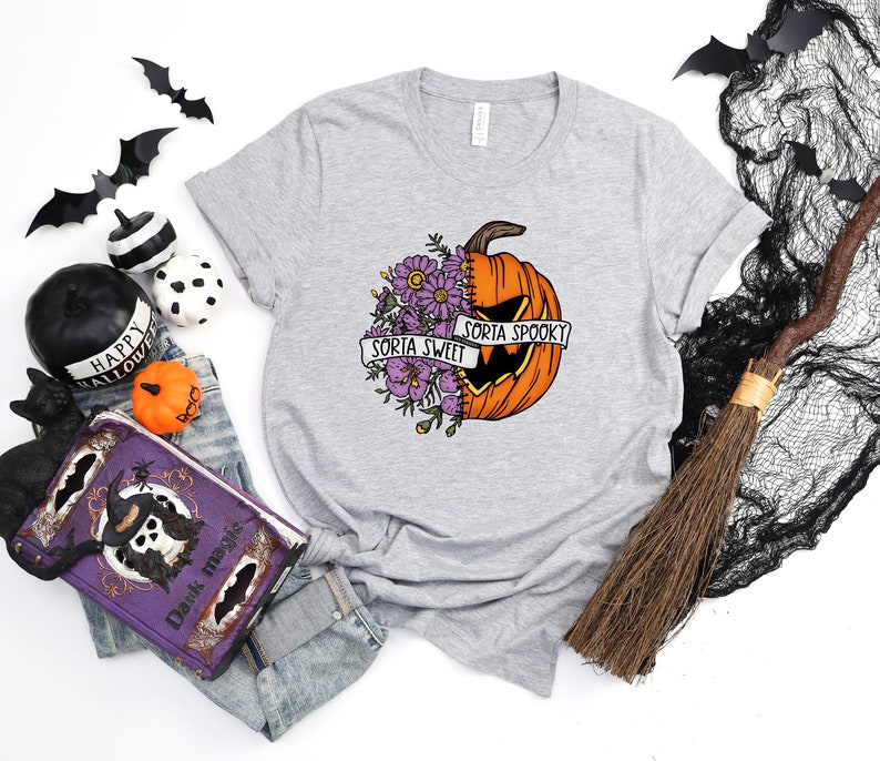 May include: A gray t-shirt with a graphic of a pumpkin with a stitched-up face. The pumpkin is half orange and half green with purple flowers on the green side. The text on the banner reads "Sorta Sweet Sorta Spooky".