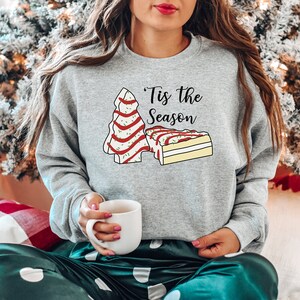 May include: A gray sweatshirt with a Christmas tree and a slice of cake design. The text on the sweatshirt reads "'Tis the Season".