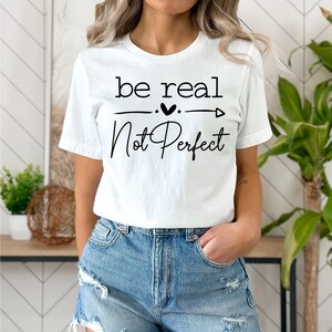 May include: White t-shirt with black text that reads "be real - not perfect" with a heart and arrow graphic.