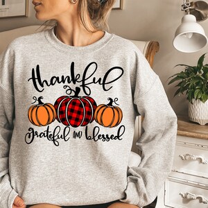 Puede incluir: Sudadera gris con un gr&aacute;fico de tem&aacute;tica de Acci&oacute;n de Gracias. El gr&aacute;fico presenta tres calabazas, una con un patr&oacute;n de cuadros rojos y negros, y el texto "thankful grateful and blessed".