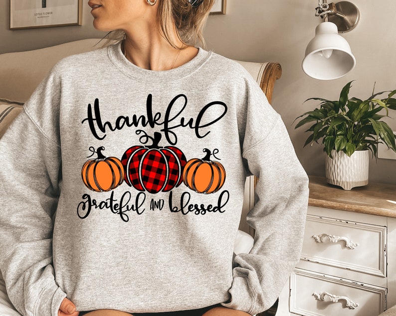 Puede incluir: Sudadera gris con un gr&aacute;fico de tem&aacute;tica de Acci&oacute;n de Gracias. El gr&aacute;fico presenta tres calabazas, una con un patr&oacute;n de cuadros rojos y negros, y el texto "thankful grateful and blessed".
