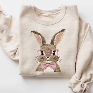 May include: A beige sweatshirt with an embroidered image of a brown bunny wearing pink glasses and a pink bow tie.