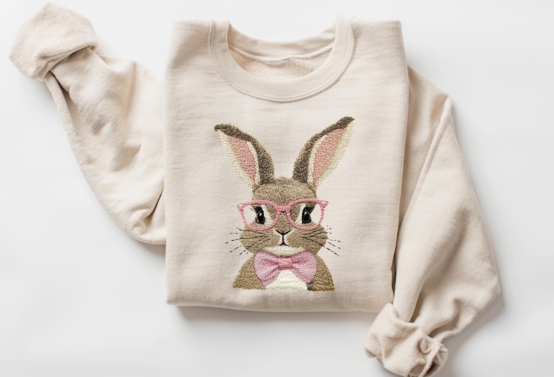 May include: A beige sweatshirt with an embroidered image of a brown bunny wearing pink glasses and a pink bow tie.