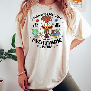 K&ouml;nnte beinhalten: Ein wei&szlig;es T-Shirt mit einer Comicfigur und dem Text "Ich habe 100 Tage &uuml;berlebt. Es ist in Ordnung. Alles ist in Ordnung." Die Figur tr&auml;gt einen Rucksack und ist von Schulutensilien umgeben.