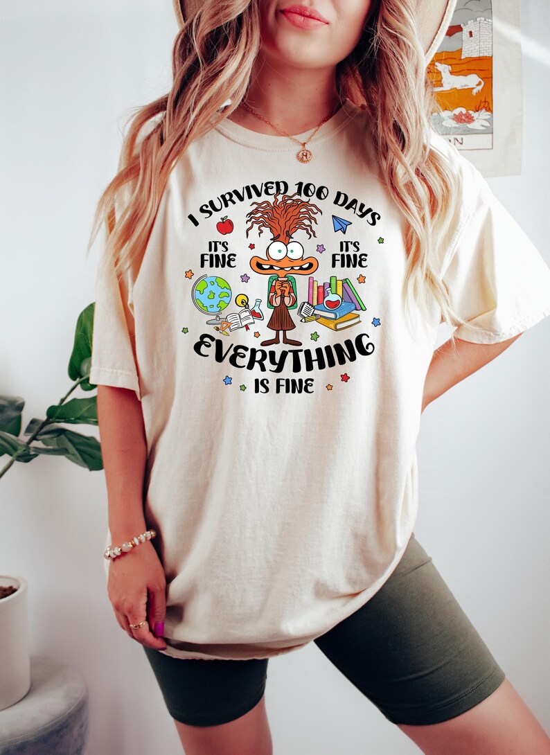 K&ouml;nnte beinhalten: Ein wei&szlig;es T-Shirt mit einer Comicfigur und dem Text "Ich habe 100 Tage &uuml;berlebt. Es ist in Ordnung. Alles ist in Ordnung." Die Figur tr&auml;gt einen Rucksack und ist von Schulutensilien umgeben.