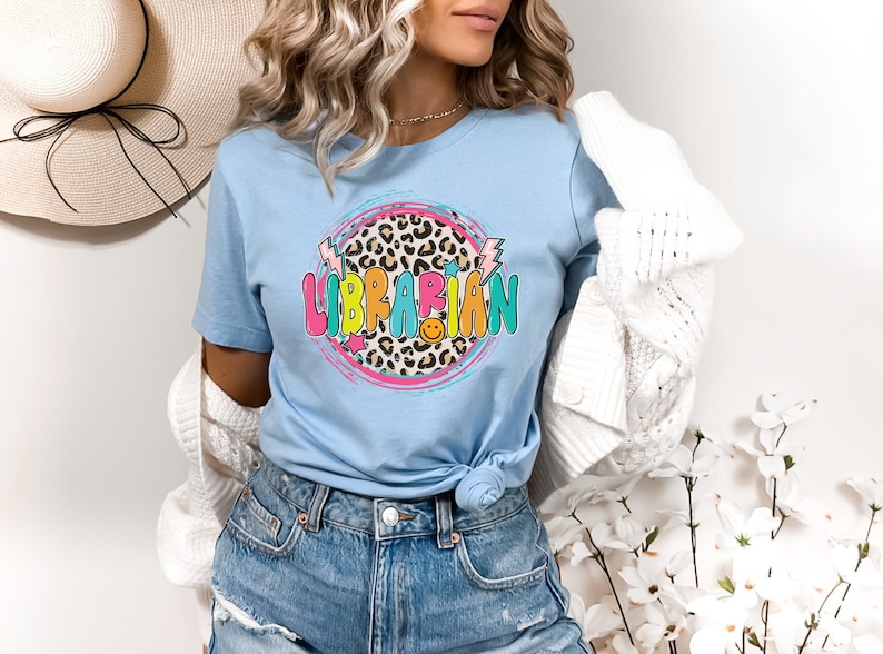 May include: A light blue t-shirt with a colorful graphic of the word "LIBRARIAN" in a circle with a leopard print background. The graphic also includes lightning bolts and stars.