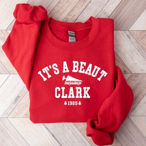 May include: Red sweatshirt with white text that reads "It's a beaut Clark 1989" with a small graphic of a car carrying a Christmas tree.