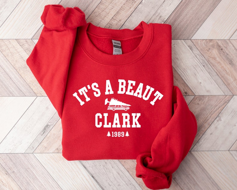 May include: Red sweatshirt with white text that reads "It's a beaut Clark 1989" with a small graphic of a car carrying a Christmas tree.