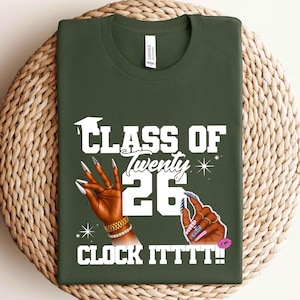 May include: Dark green t-shirt with the text "CLASS OF Twenty 26 CLOCK ITTTT!!" in white. The design includes illustrations of hands with long, manicured nails, a graduation cap, and a pink lip.