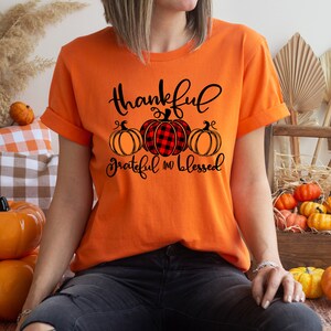 Puede incluir: Camiseta naranja con un gr&aacute;fico negro de tres calabazas. Las calabazas est&aacute;n en fila y la calabaza de arriba es de cuadros rojos y negros. El texto en la camiseta dice "Thankful Grateful and Blessed".