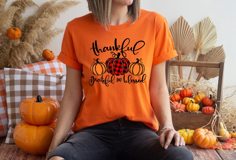 Puede incluir: Camiseta naranja con un gr&aacute;fico negro de tres calabazas. Las calabazas est&aacute;n en fila y la calabaza de arriba es de cuadros rojos y negros. El texto en la camiseta dice "Thankful Grateful and Blessed".