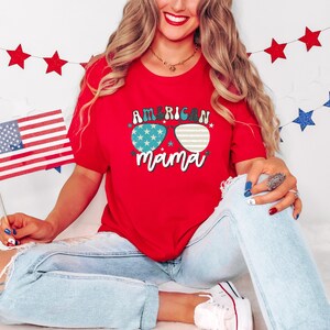 Puede incluir: Camiseta roja con un gr&aacute;fico de gafas de sol con estrellas y el texto "American Mama". La modelo lleva pantalones vaqueros azules y sostiene una peque&ntilde;a bandera estadounidense.