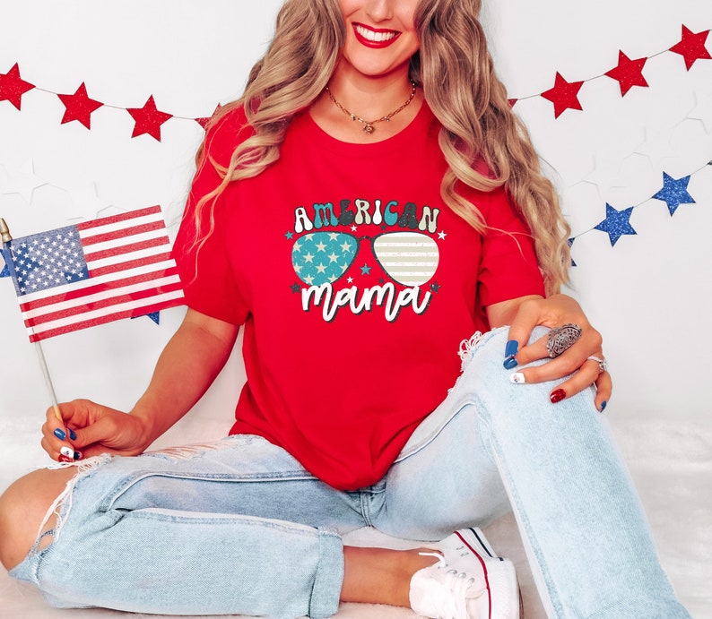 Puede incluir: Camiseta roja con un gr&aacute;fico de gafas de sol con estrellas y el texto "American Mama". La modelo lleva pantalones vaqueros azules y sostiene una peque&ntilde;a bandera estadounidense.