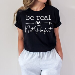 May include: Black t-shirt with white text that reads "be real. Not Perfect" with a small heart and arrow graphic.