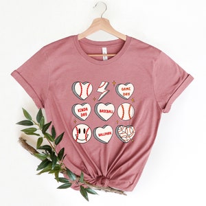 May include: A dusty rose t-shirt with a graphic design of baseballs in hearts with phrases like "Game Day", "Kinda Day", "Baseball", "Ballpark".