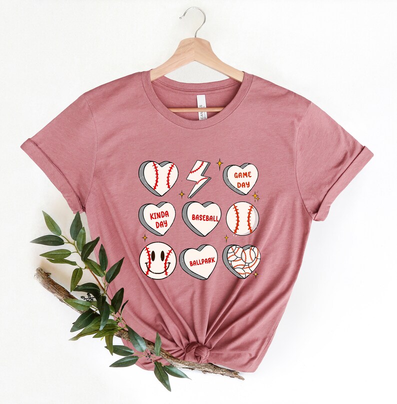 May include: A dusty rose t-shirt with a graphic design of baseballs in hearts with phrases like "Game Day", "Kinda Day", "Baseball", "Ballpark".
